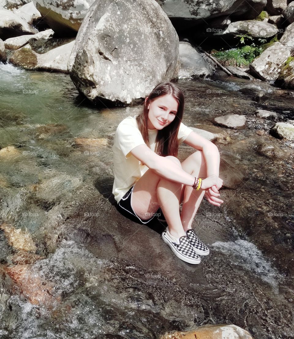 Capture of beautiful girl in nature, perched on rock with clear river running.