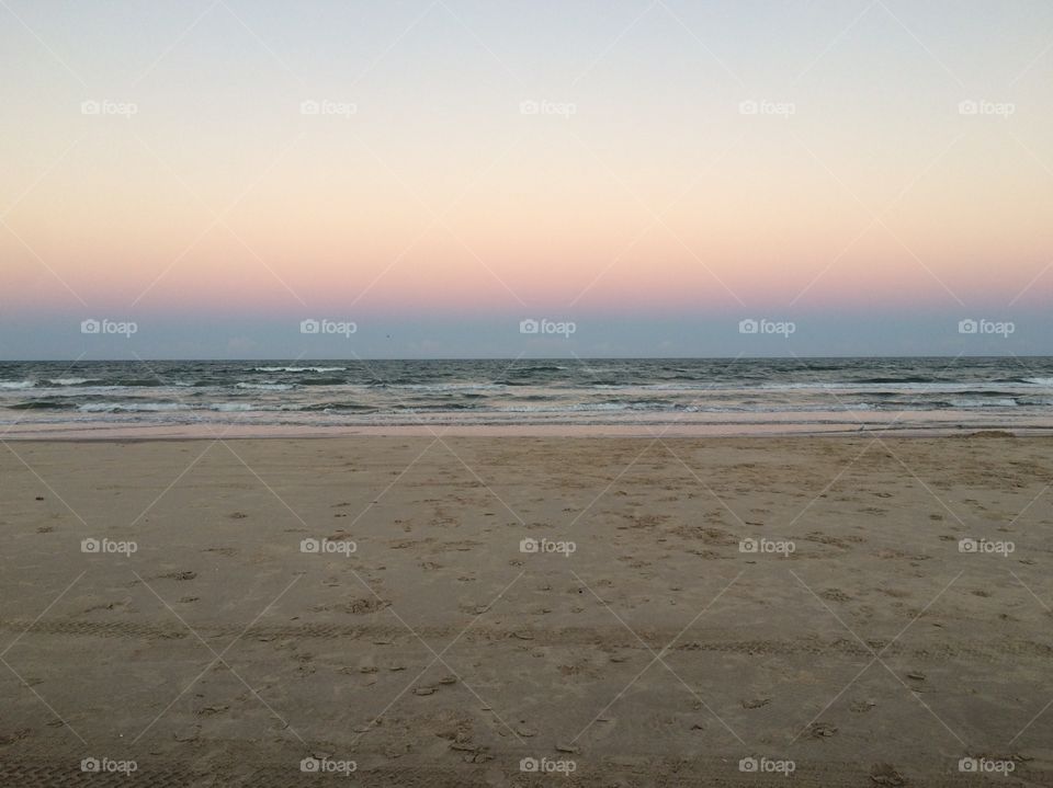 A horizontal image of the seashore. The sun is setting, though the sun is not shown. The sky has yellow, pink, blue, and purple tones. The waves meet the sky at the horizon.