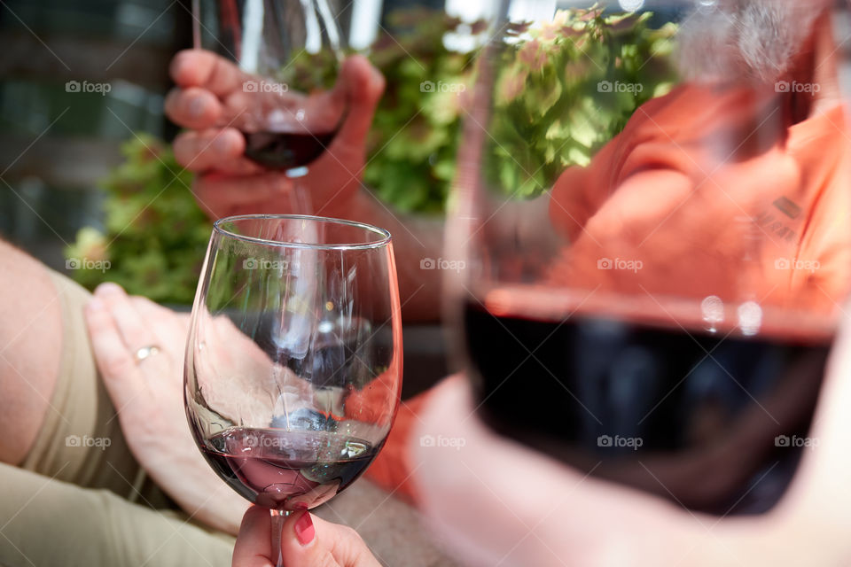 Three people sitting on a deck in the sunlight all holding wine glasses. A close shot of their hands, with the center glass being in sharp focus and the background and foreground are blurred.
