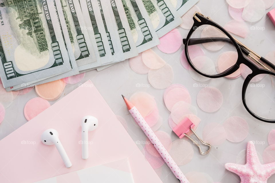 Flatlay items: cash money one hundred dollars bill, reading glasses, wireless headphones, notebook, pencil, star pink color, confetti lying on gray concrete background