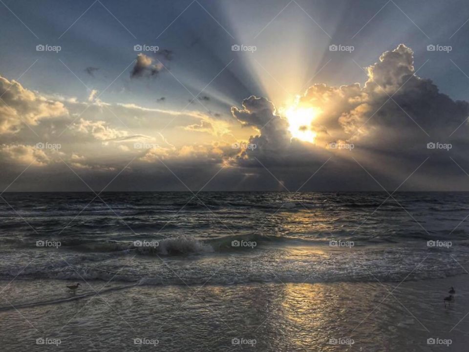 Rays of sunlight shining across the sky from behind the clouds and reflecting on the waves of the ocean at sunset 