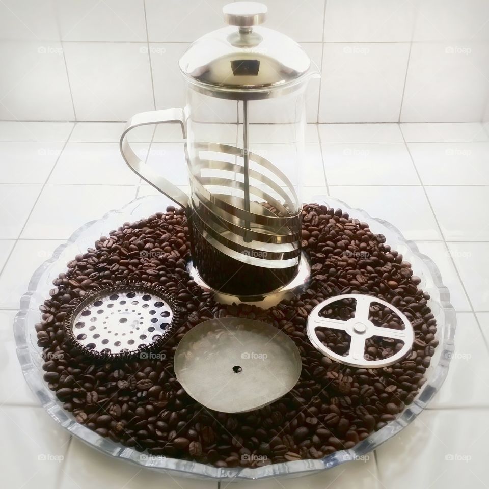 A french press coffee maker with the strainer pulled apart sitting on whole coffee beans on a round glass tray in a white tile kitchen in early morning