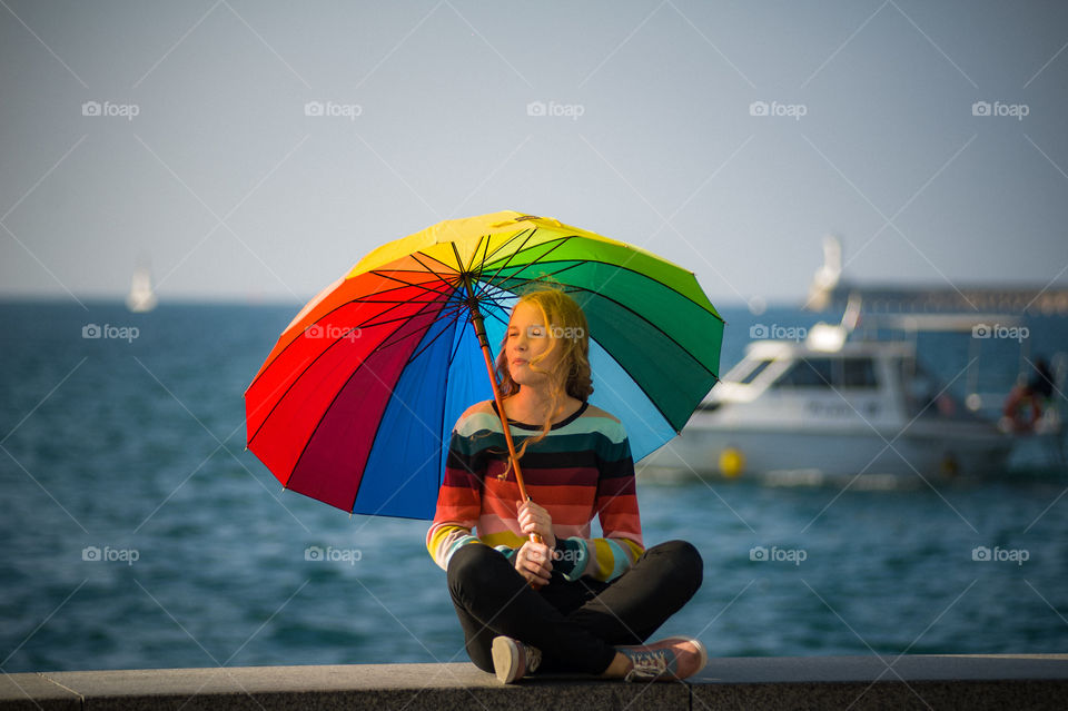 young girl sits near the seashore with bright multi-colored umbrella