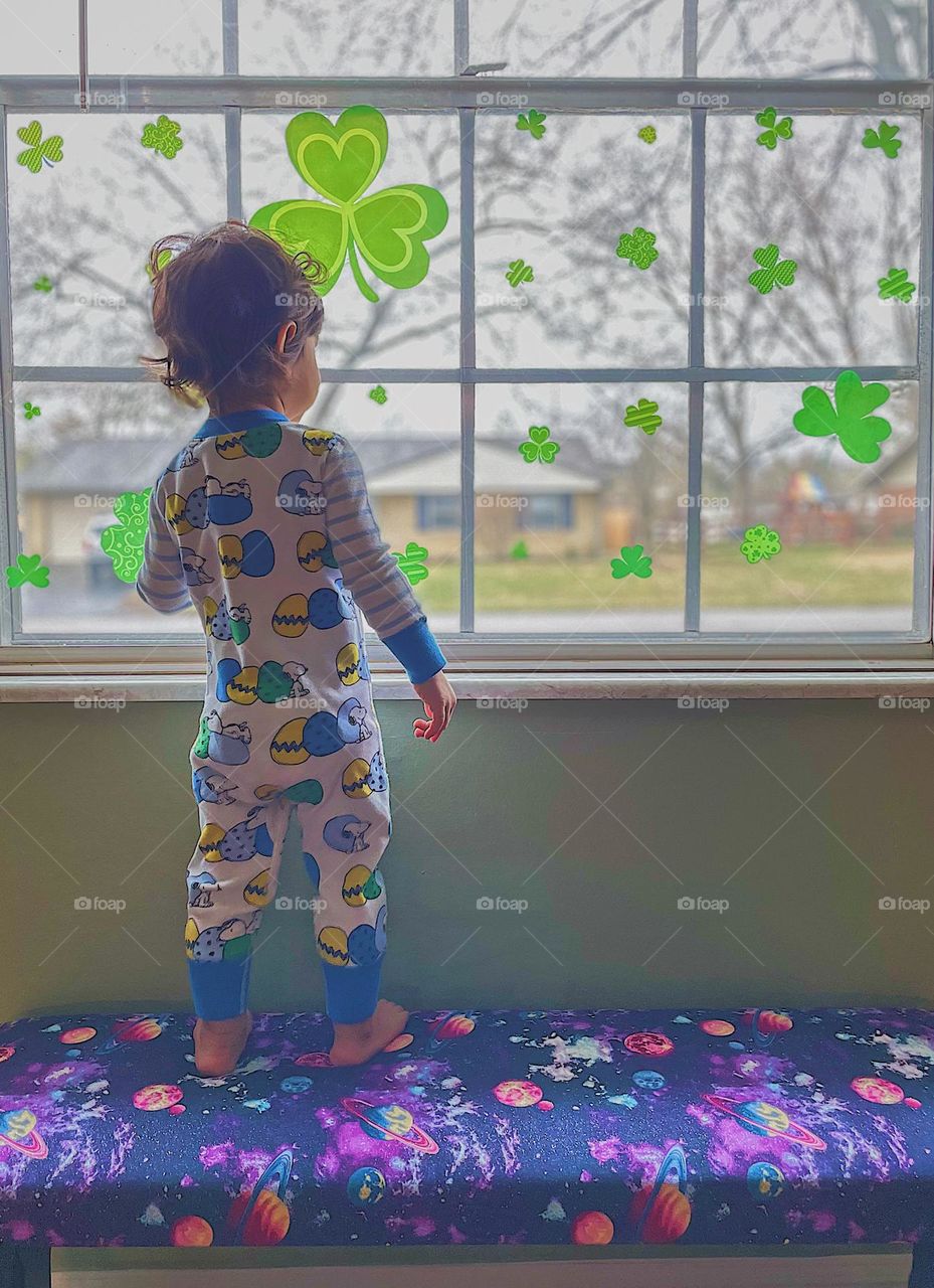 Toddler girl looking out window, toddler is looking at neighbors, toddler girl dressed in Easter outfit looks out window decorated with shamrocks, celebrating all the holidays, toddler girl at the window, toddler girl on neighborhood watch 