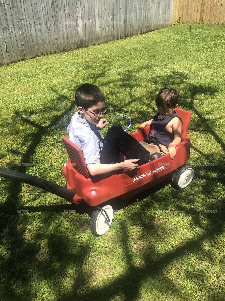 Two boys aged 10 and 2 1/2  that are cousins, sitting in a red wagon playing together  on the green lawn. 