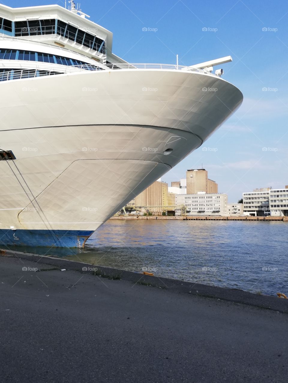 A cruiser is on its berth in the port terminal. The bow of the passenger ship is white and wide, the bottom is blue. Across the water there is a dock and some buildings and cars.