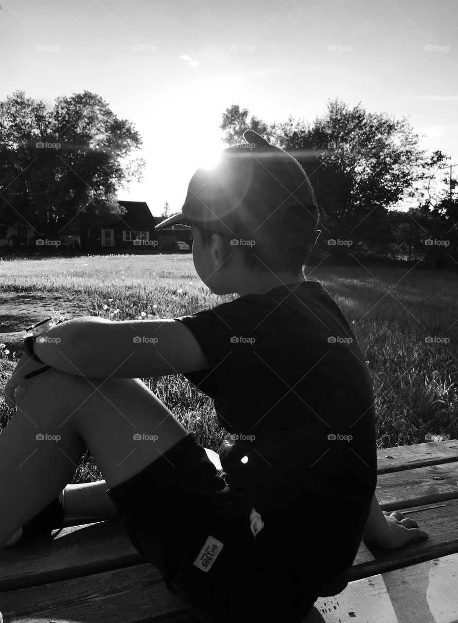 Boy looking out into the distance sitting on a park bench with the sun shining down