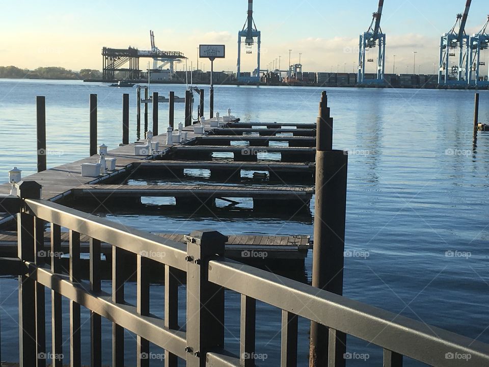 Angled railing, empty boat  dock in blue water with industrial cranes on opposite shore