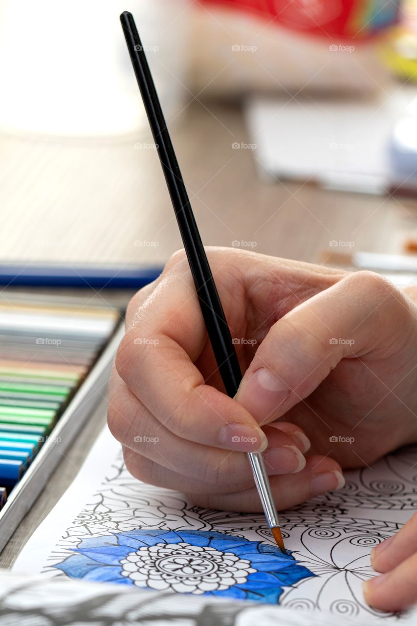 A portrait of a person painting blue with a thin paint brush in a coloring book for adults. only the hand is visible. in the back there is a colorful case of multiple colored pencils.