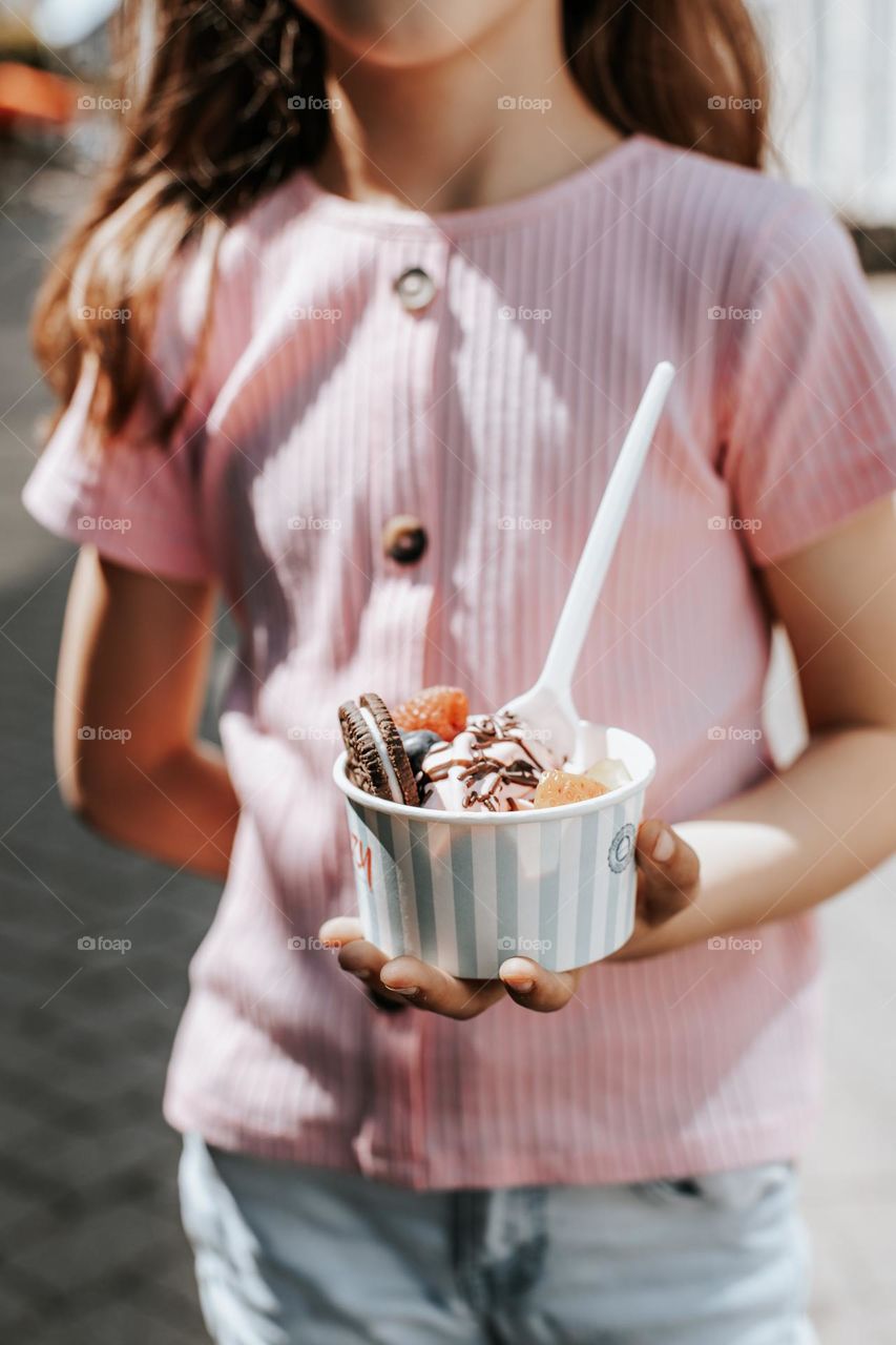 Portrait of one caucasian unrecognizable girl in a pink t-shirt holding a paper cup with ice cream decorated with cookies, berries and chocolate sauce, standing on a city street on a summer sunny day, vtd side close up.