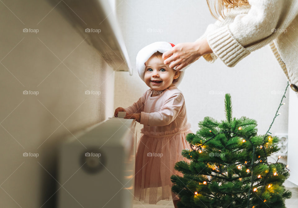 Cute baby girl in Santa hat with her mother in room with Christmas tree at home 