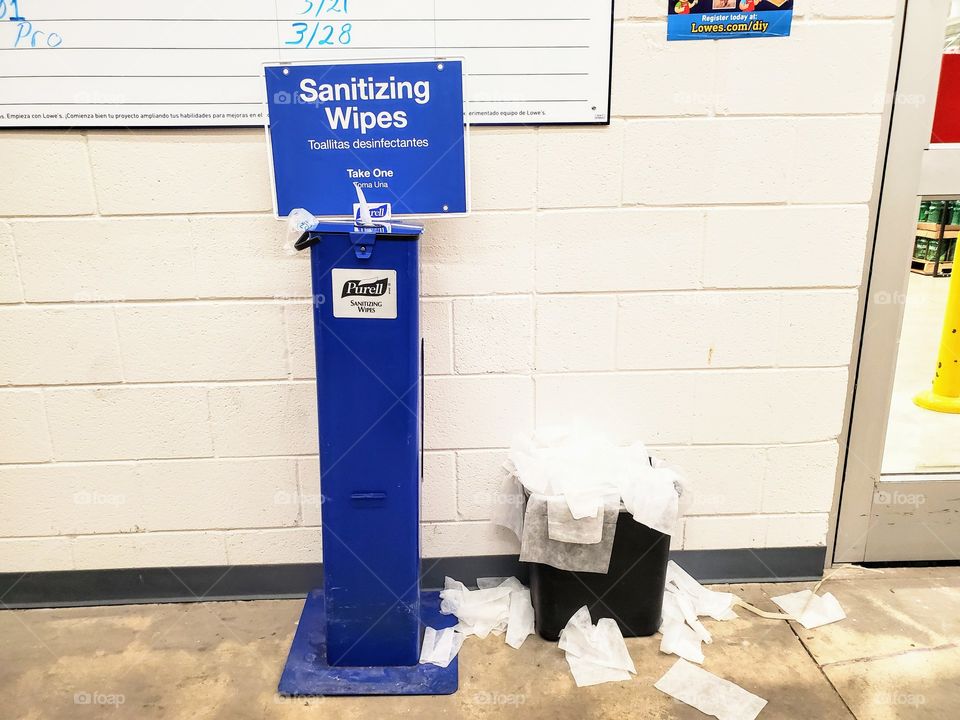 Blue sanitizing wipes station next to an overflowing trash bin at a retail store during the Corona virus world pandemic.