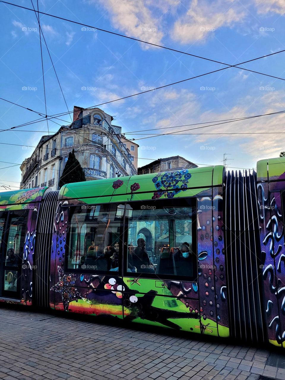 Multicolored tramway travelling in front of chic building under sunny weather and electric wires.