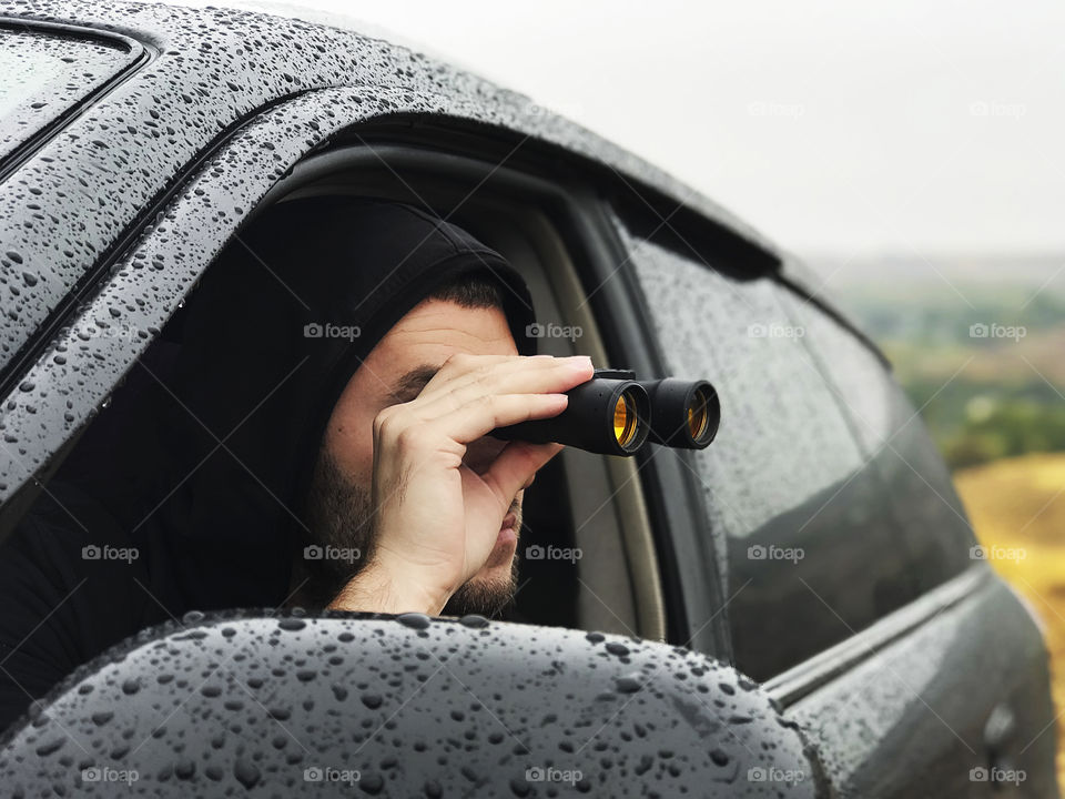 Young man watching through binoculars out of wet cat at rainy day 