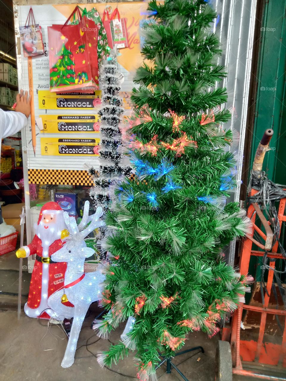 CHRISTMAS TREE ON BROAD DAYLIGHT,, SERIES LIGHTS ARE STILL ON WITH DWARF SANTA CLAUS,, IN FRONT OF A STORE