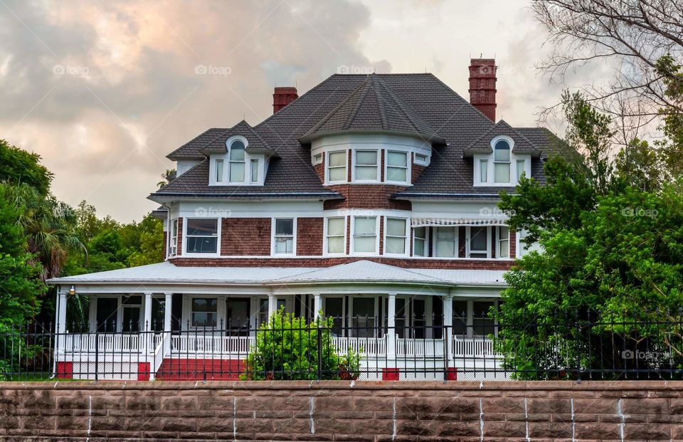 beautiful big vintage historic house with red brick, white trim, and a large wrap around porch with sky of clouds reflecting the orange sunset