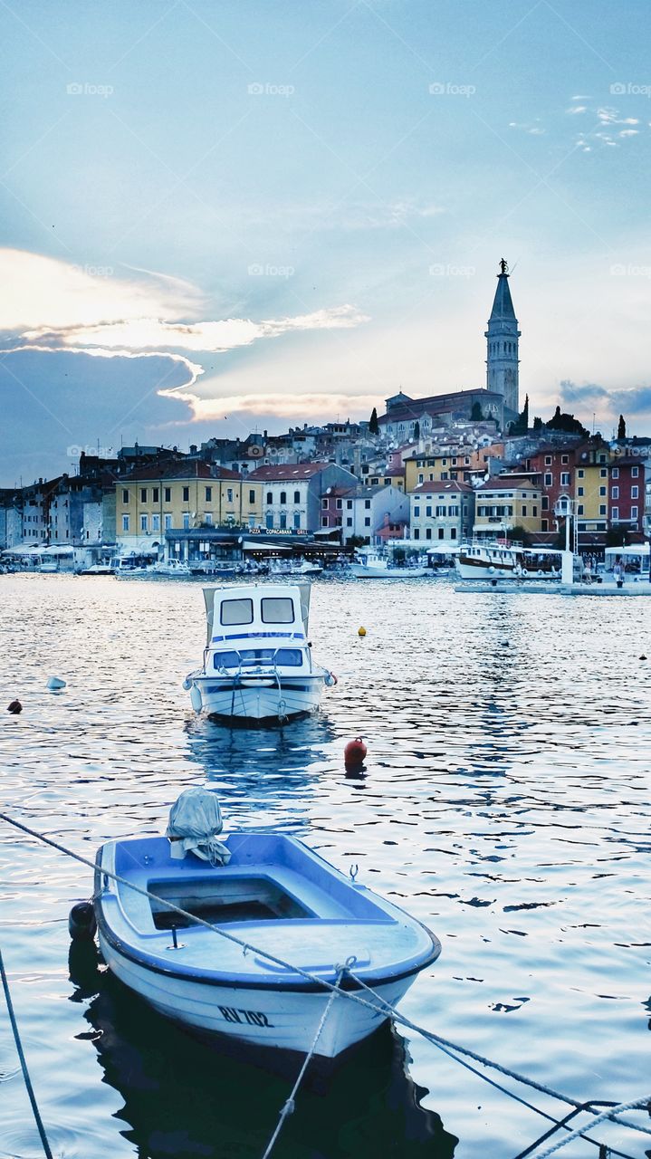 The tower at the church of St Euphemia stands over the city of Rovinj, Croatia, behind the sea, harbour or boats at dusk.