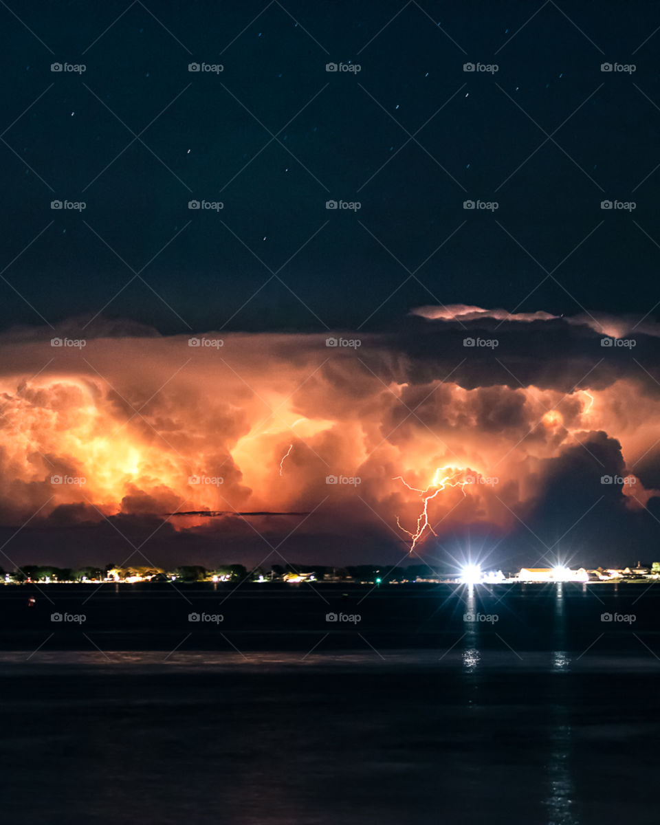 Lightning illuminating low thunderstorm clouds far off shore, with a starry night sky above the storm. 