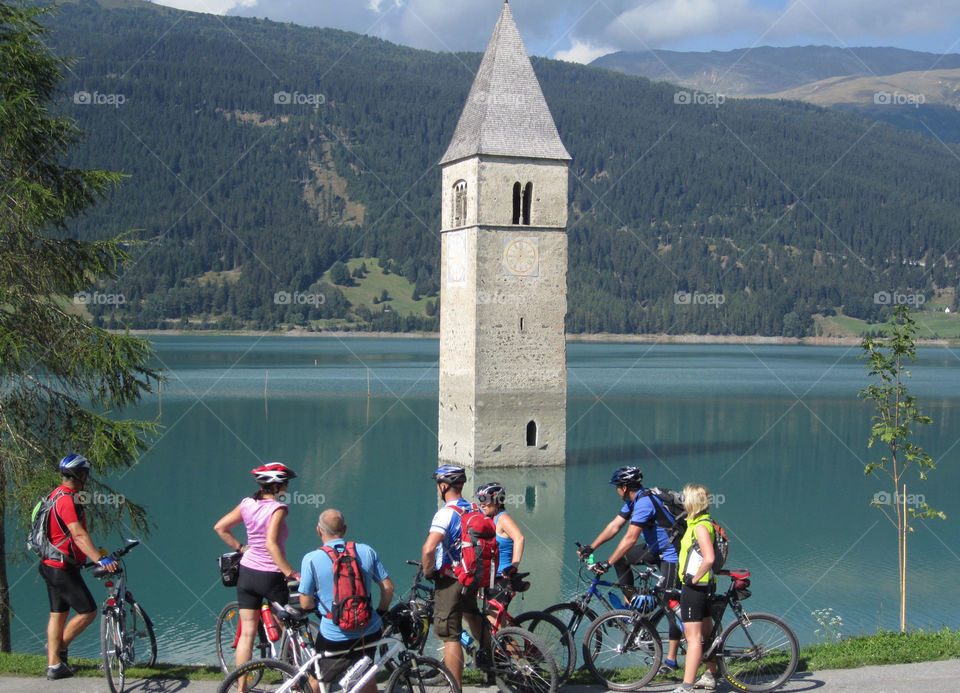 Travel in the Alps by Bike. Cyclists admire the church tower in Lake Reschen. Lake Reschen is the largest lake above 1,000 metres in the Alps.Located south of the Reschen Pass, in South Tyrol, Italy the area is popular with cyclists.