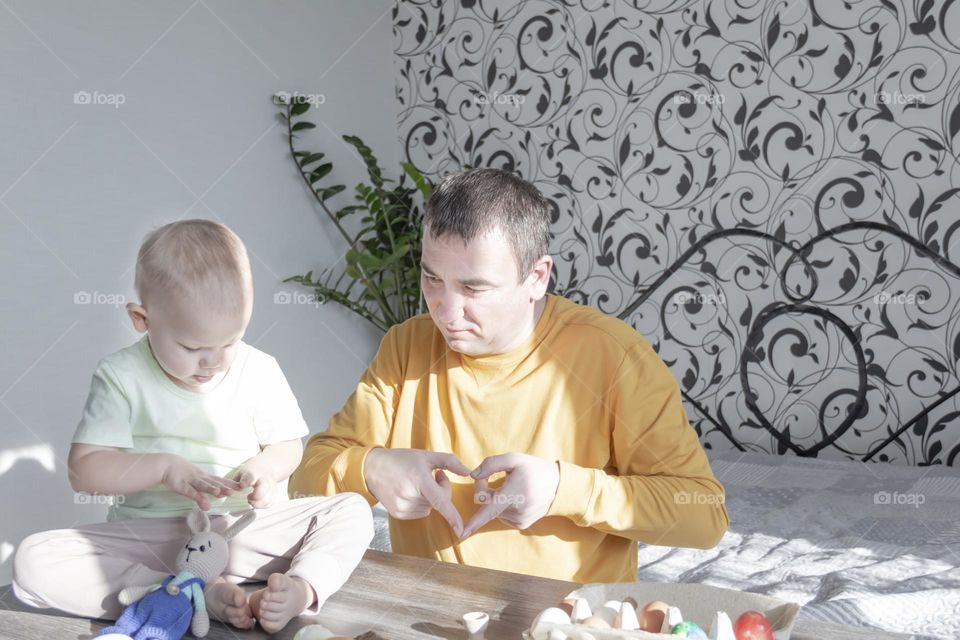 A dad and a little boy prepare for Easter at home indoors, paint Easter eggs with early-colored paints, sit at a table, sunlight falls on a wooden table.