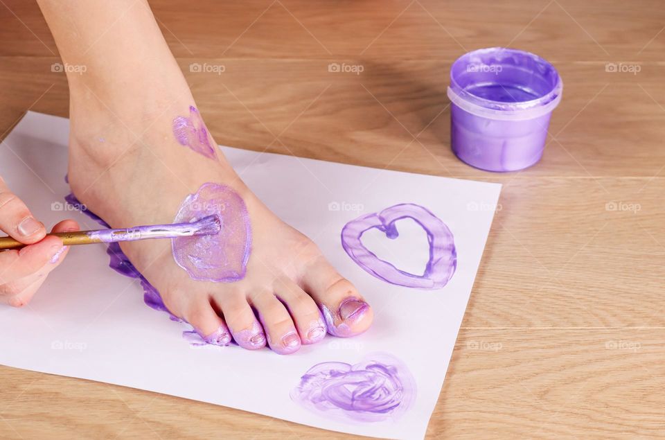 Caucasian teenager girl paints her foot holding on a white sheet of paper with lilac paint with a brush, sitting on a wooden floor, close-up side view. Kids creativity concept.