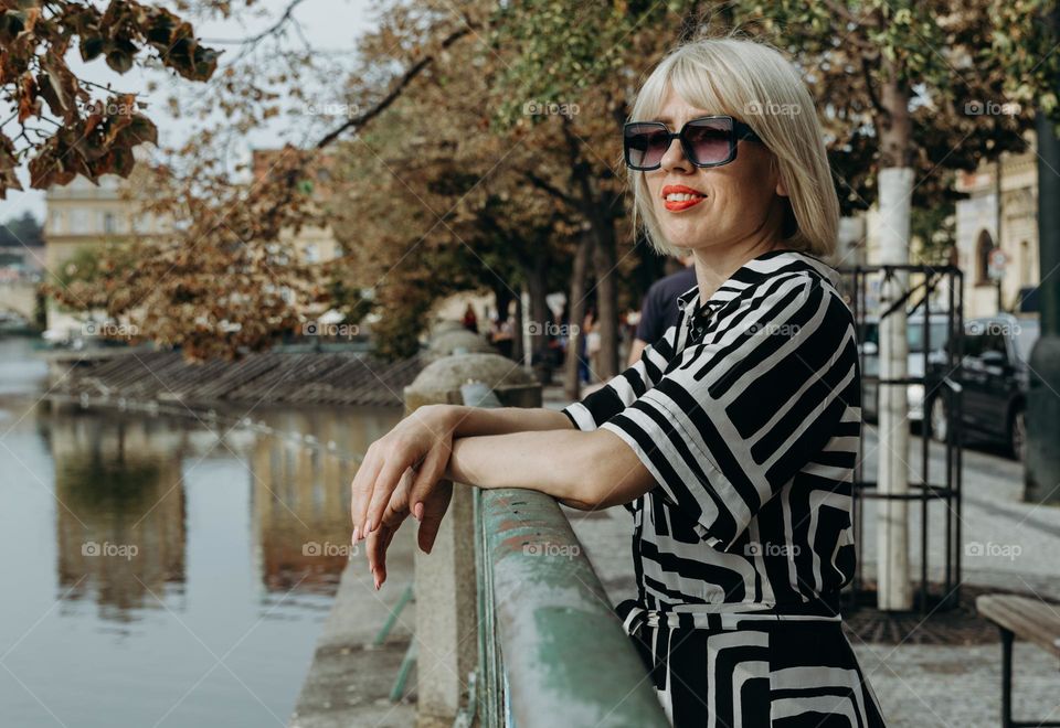 Portrait of a young beautiful blonde Caucasian girl in sunglasses standing on the bridge of the city embankment, resting her hands on the railing and looking with a smile in the distance on a sunny autumn day, close-up view from the side.
