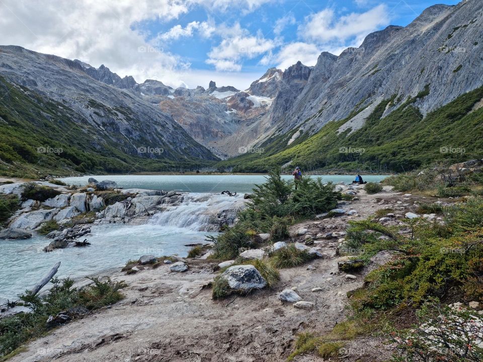 turquoise water lagoon that descends from a glacier on top of the mountain, surrounded by mountains with green vegetation and sky with clouds.