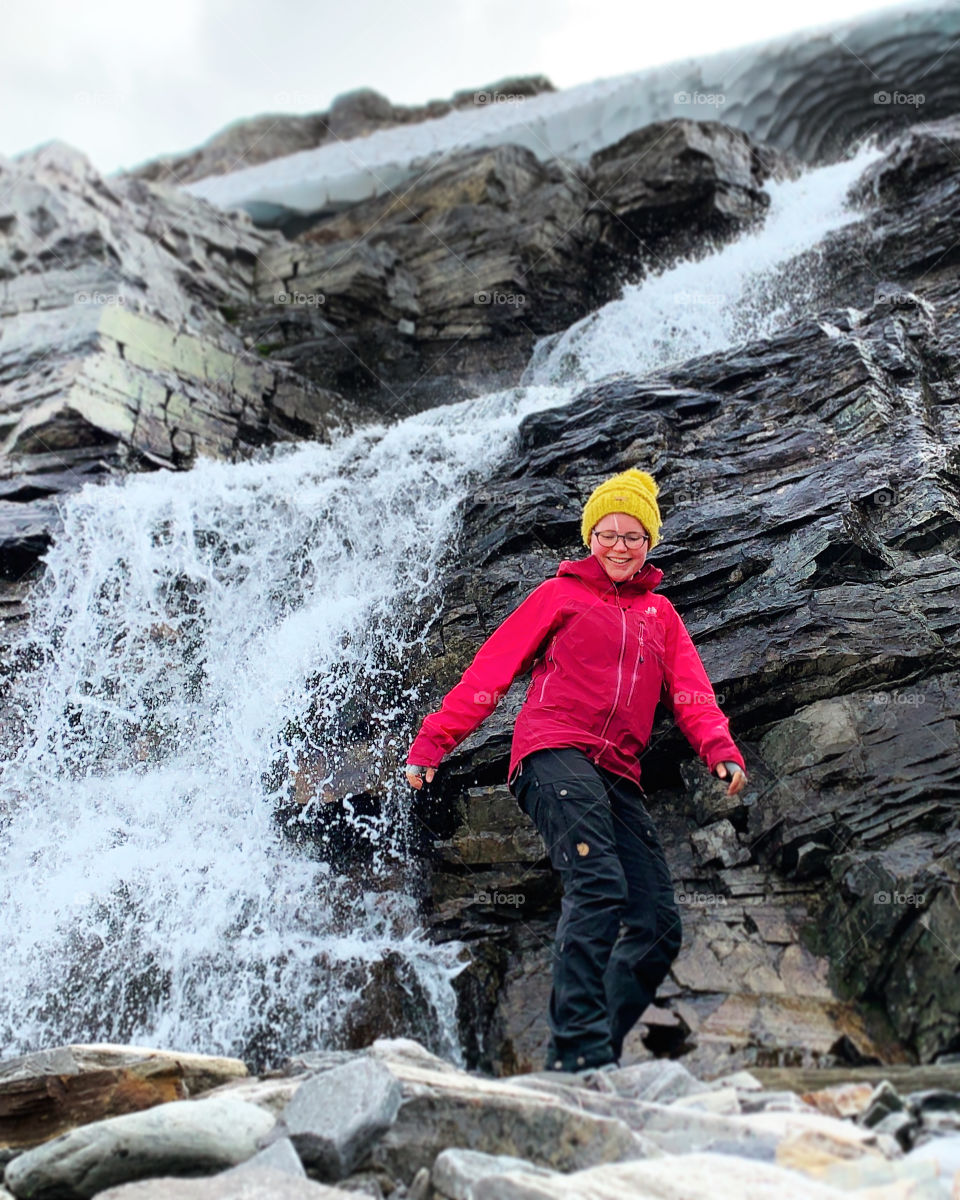 Female hiker exploring a beautiful waterfall. Smiling girl wearing a red jacket balancing on rocks. Solo outdoor adventure in the wild and remote Finnish Lapland. A moment of happiness while hiking the Nordkalottleden trail.