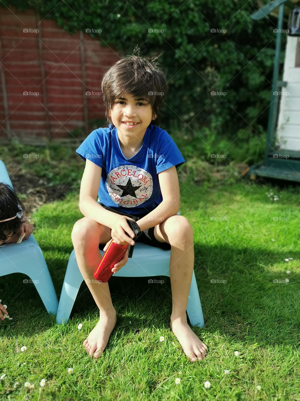 Barefoot shaggy haired boy sitting in the garden with sprat an in hand
