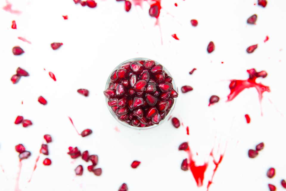 Glass bowl filled with red fresh pomegranate seeds. Flat lay on a white background with scattered pomegranate seeds and juice around. Fresh summer fruit for a healthy lifestyle.