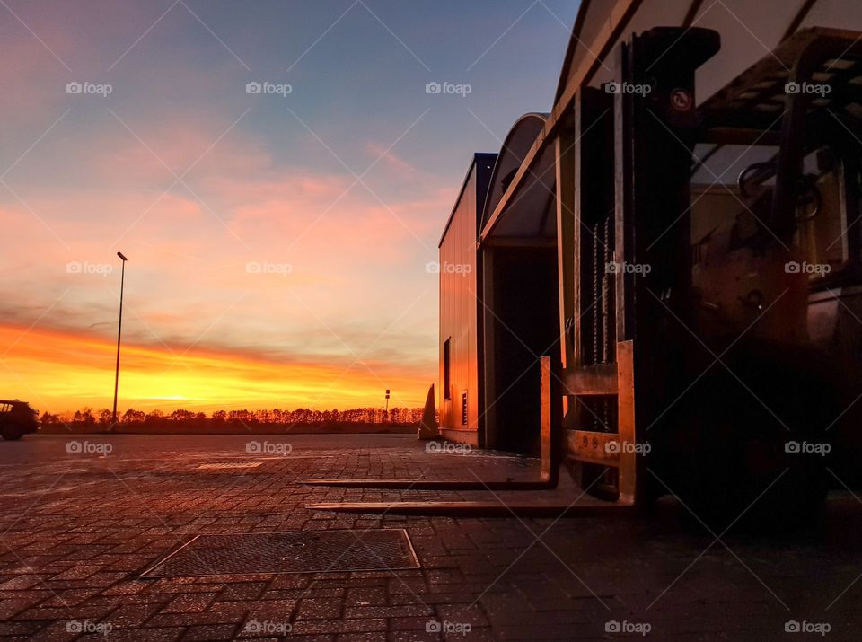 A magical and scenic sunset at an industrial factory making it almost completely orange. in the foreground there is a fork lift parked in a parking lot.