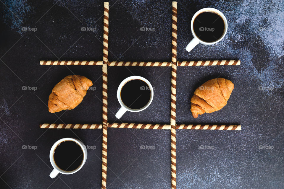 Working morning with cups of hot black coffee and sweet croissant on black background. Tic tac toe game. Top view. Flat lay. Creative concept of modern breakfast food.