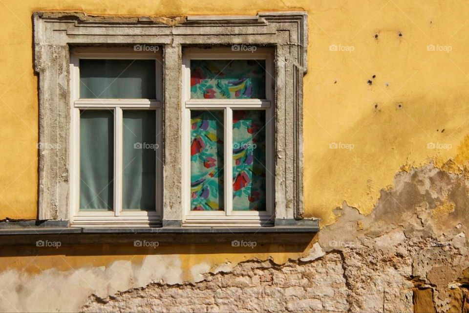 A weathered yellow house facade with an old window and colorful curtains