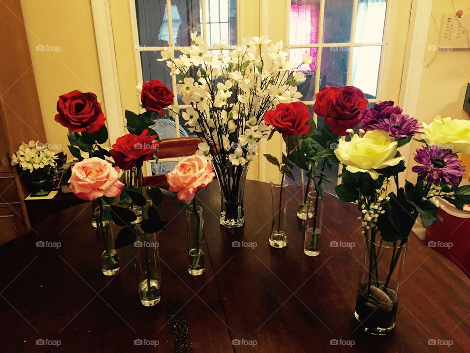 A bevy of beautiful flowers, on a table top, including several long stemmed red roses in single vases, pink roses, large vase full of white dogwood blossoms, a purple and lavender, white rose arrangement in a vase. 