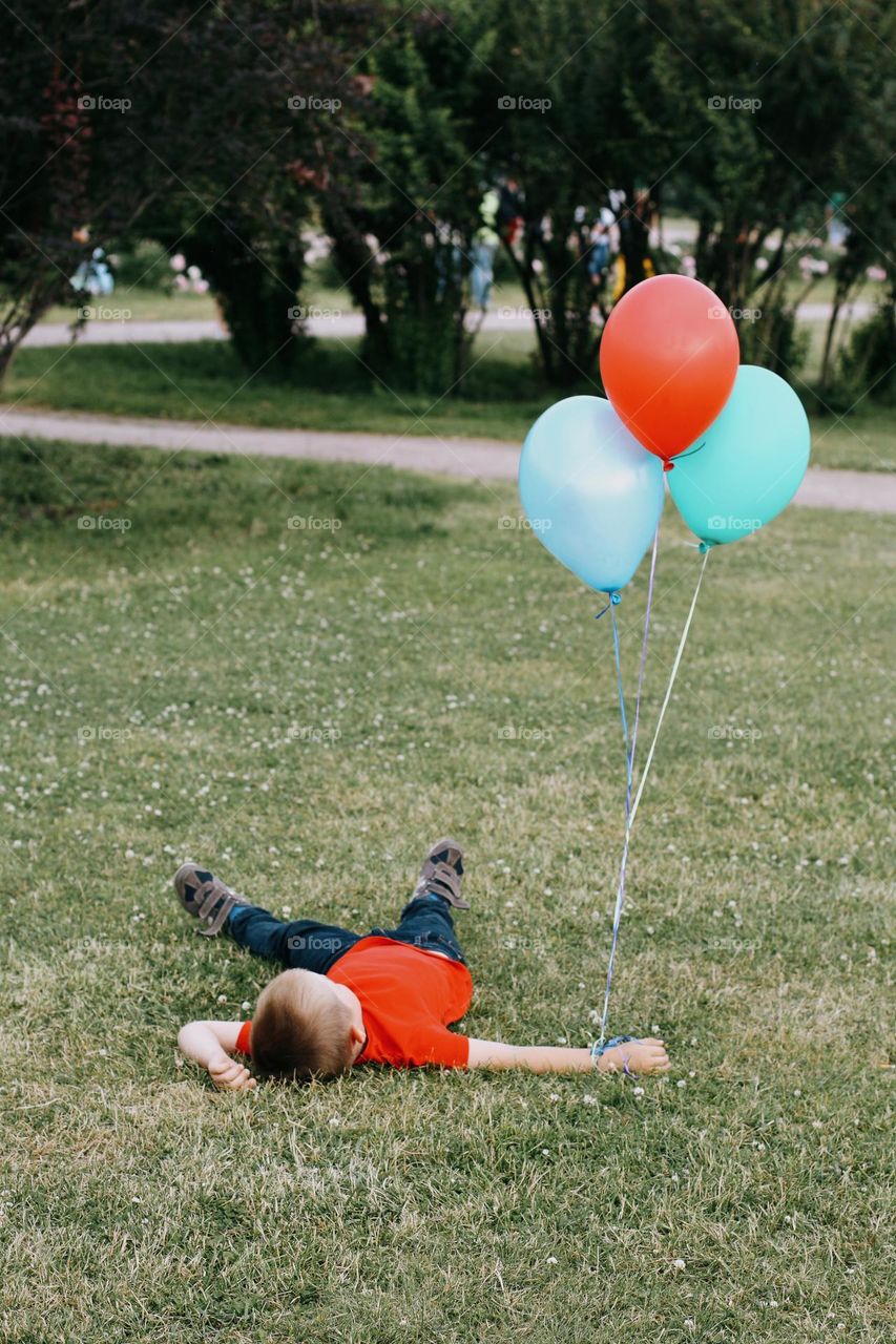 A little blond boy in a red T-shirt lies on a green field, holding a bunch of colorful balloons in his hand, back view 