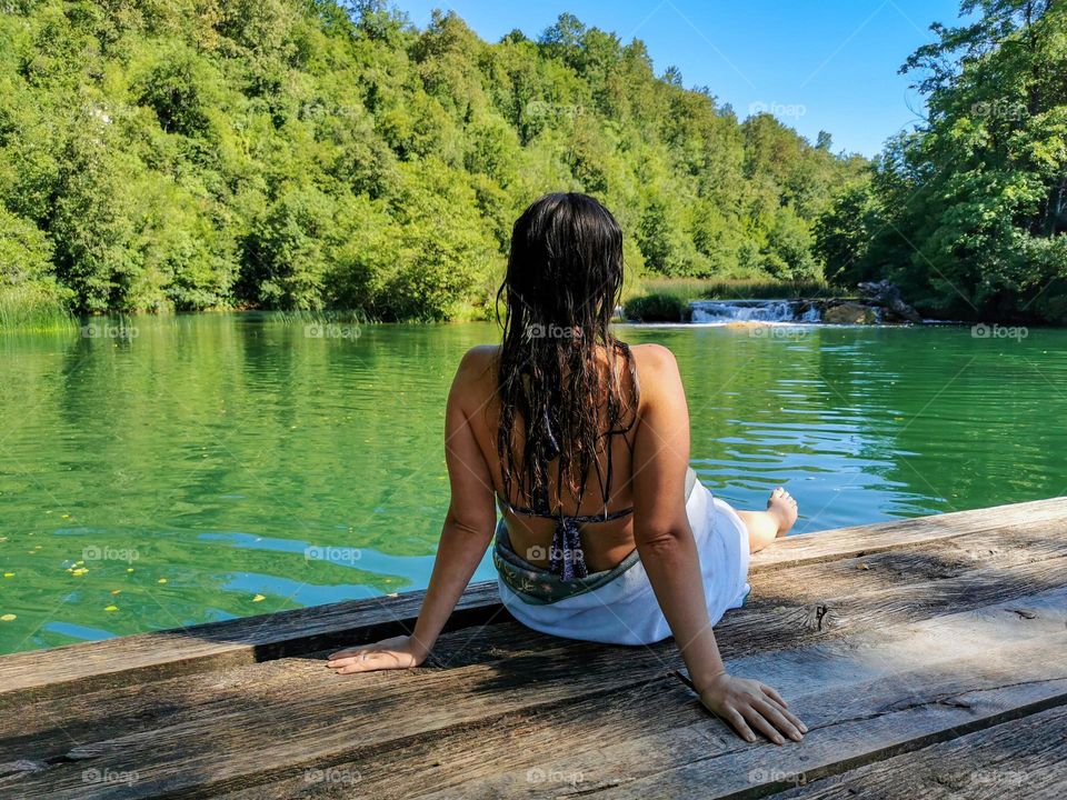 Rear view of young woman wrapped in a towel sitting on wooden pier by a beautiful green river on a sunny day in summer