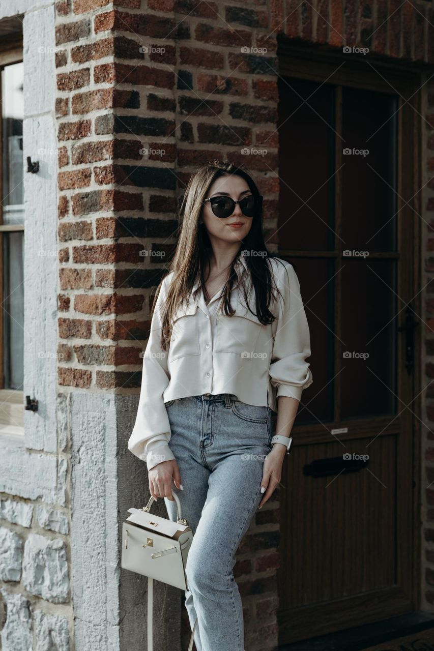 A portrait of one beautiful young Caucasian brunette girl in sunglasses, loose long hair, in stylish and fashionable clothes stands near her house, getting ready to go for a walk on a spring sunny day, close-up side view.