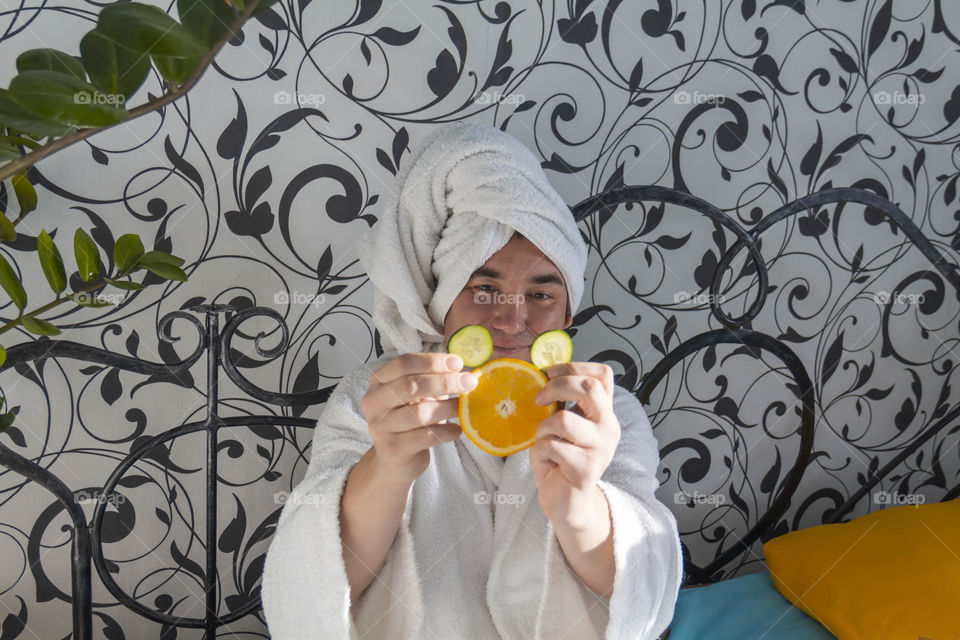 A man and home spa care at home takes care of himself and smears his face with cream, does a manicure at home on a blue background and with oranges and rests.