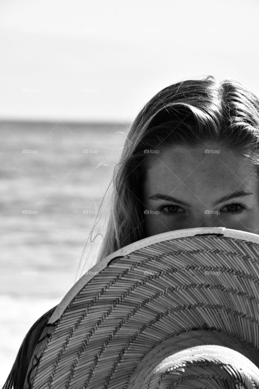 black and white portrait of girl at beach covering face with hat only eyes