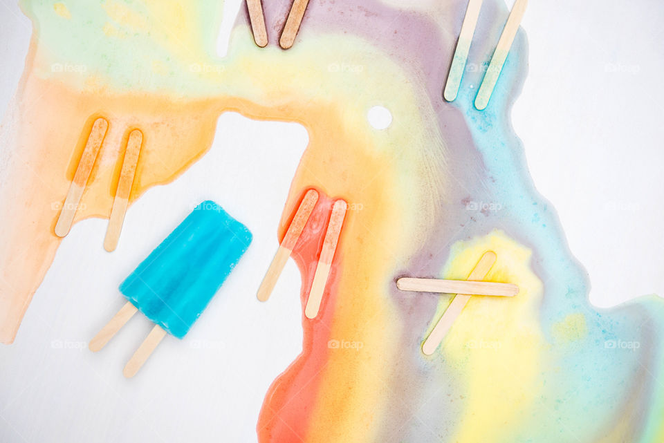 Flat lay of a bright blue frozen popsicle surrounded by multicolored melted popsicles with popsicle sticks and mixed colors remaining