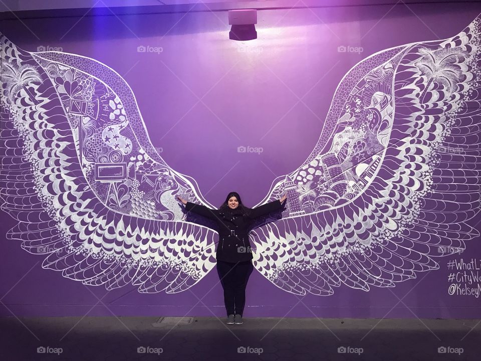 Girl with wings ready to fly. Arms wide open in front of a purple background.