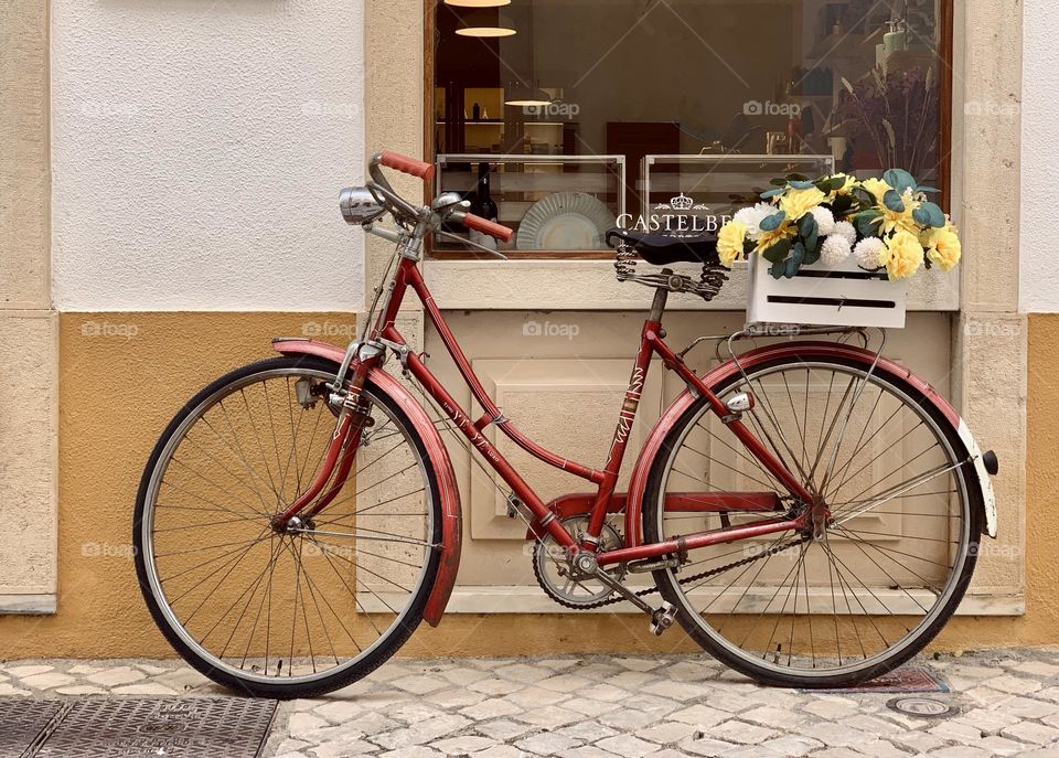 A vintage red bicycle propped up outside a shop, with a wooden crate of flowers on the back