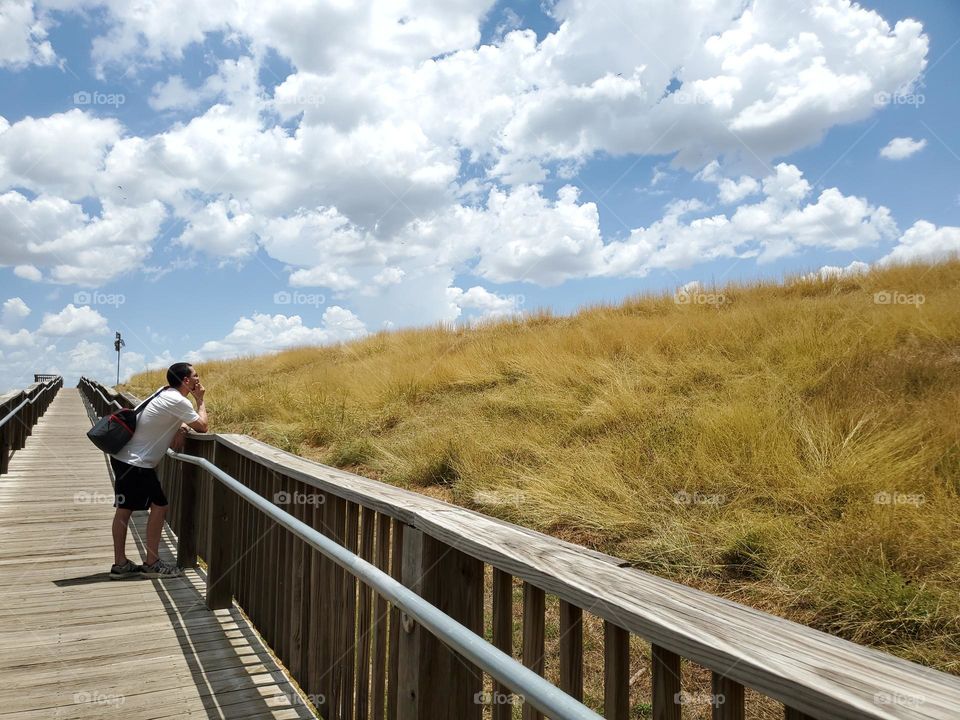 A man leaning on the rails of a wood deck path as he peacefully gazes out to the clouds beyond the golden grassy hill. He is alone and in a moment of stillness that transcends to meditative state that nature often brings.