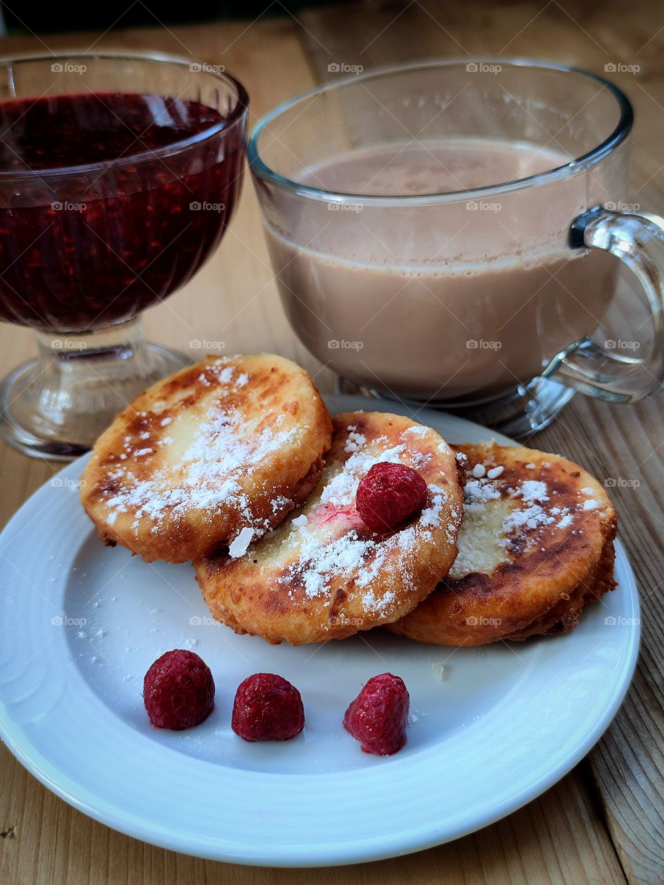 Breakfast. On a plate there are three cheesecakes sprinkled with powdered sugar and raspberries. Next to the plate is a vase of raspberry jam and a cup of hot cocoa