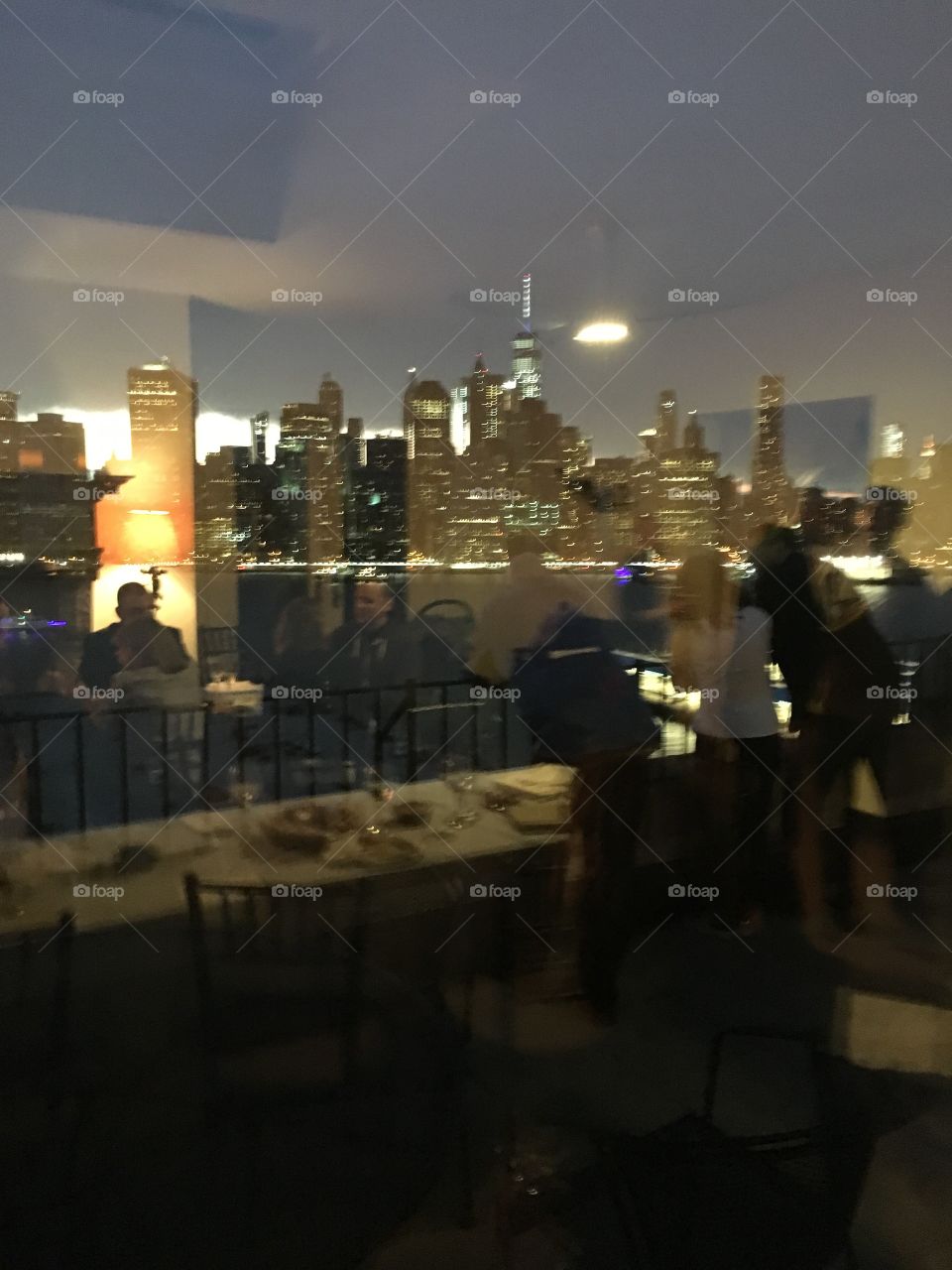 An elite home at Pierrepoint Place Brooklyn, NY in a high rise with large windows, balconies and views of Manhattan and Brooklyn all around. Manhattan is reflecting in the window while the dinner guests can be seen through the glass. 2017 Hypnotic Pr