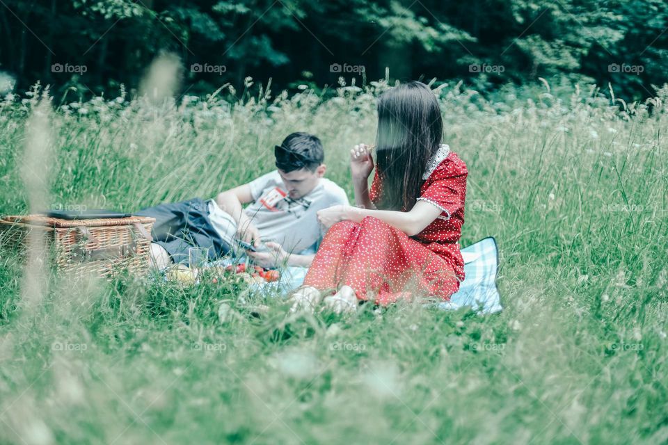 Young beautiful caucasian couple of a guy with a phone in his hands and a girl in a red polka-dot dress eating fruit romantically resting on a picnic in the forest, close-up side view. The concept of family picnic, romantic couple, outdoor recreation