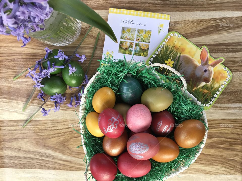 Easter eggs in a basket, on a wooden table, surrounded by the easter card and purple hyacinth flowers in a wase.