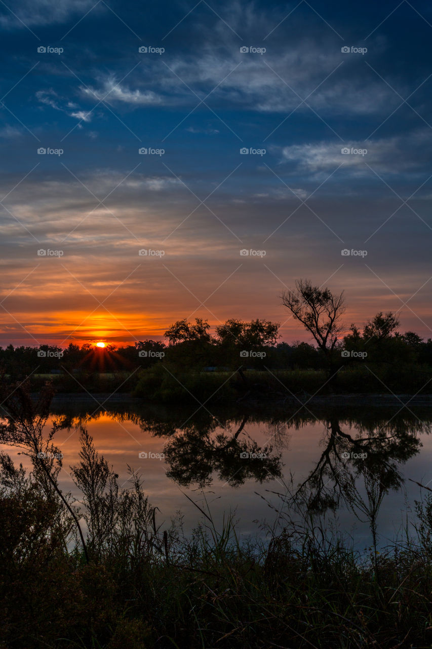 The early Spring sun rise with vivid red and orange colors is reflected in a stock pond with silhouettes of distant trees