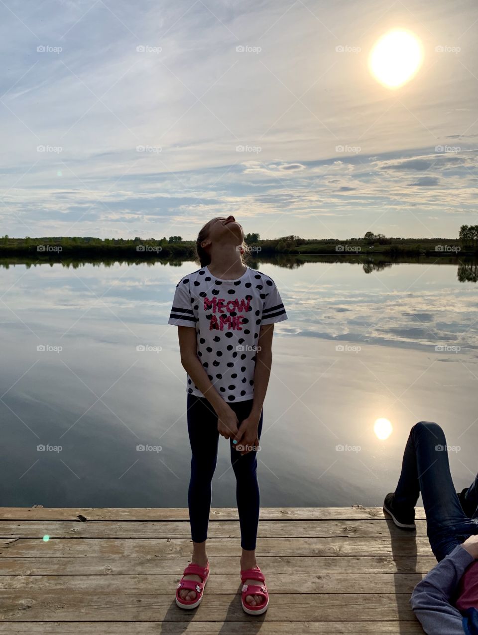 girl on the lake looks at the sky and enjoys the sunset. Water reflects the sky, clouds and the sun