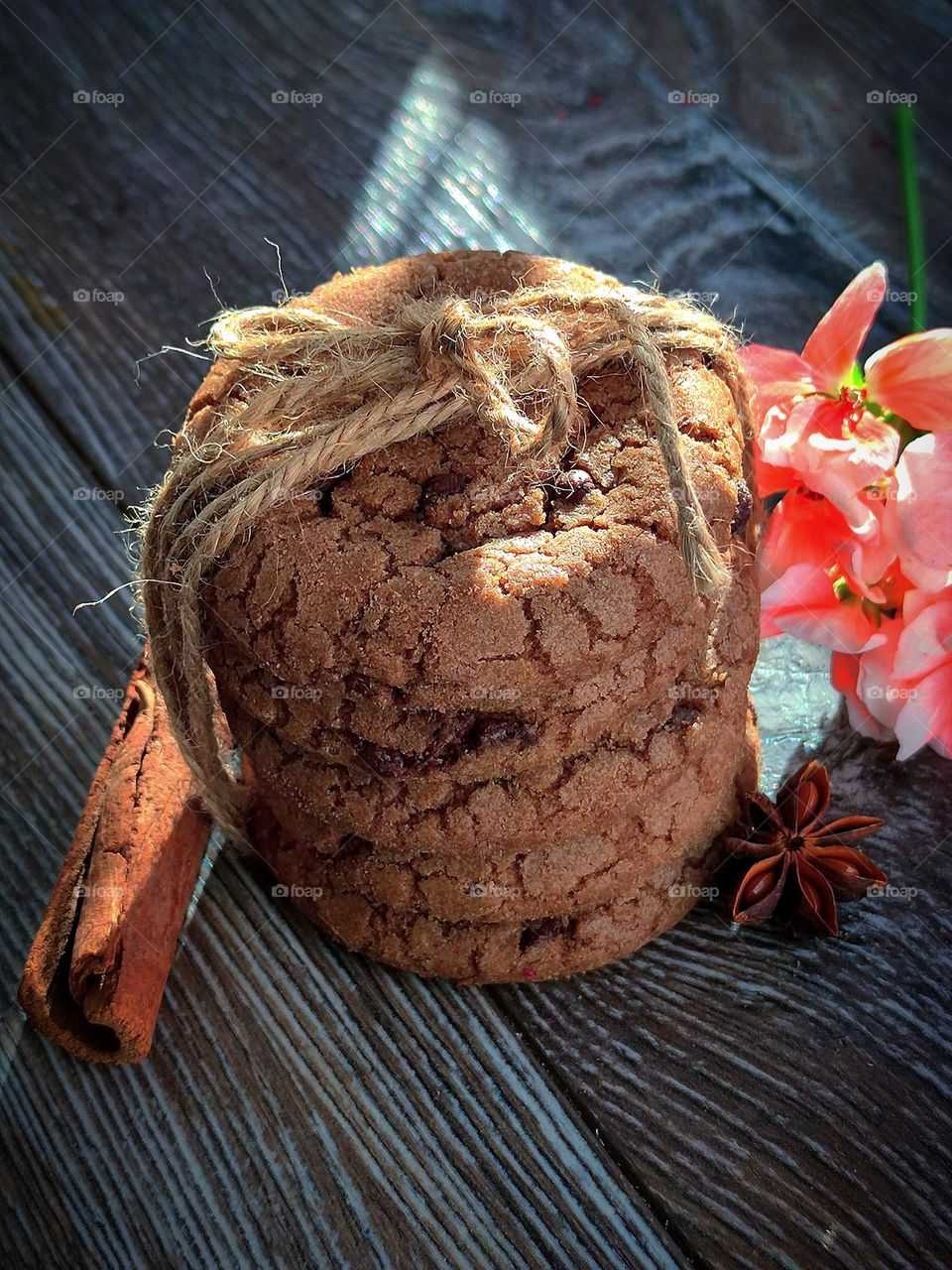 Chocolate chip cookies tied with gray twine.  Nearby, on a wooden background are: cinnamon stick, star anise fruit and pink geranium flowers.  A ray of the sun falls on objects, which gives contrast and energy.